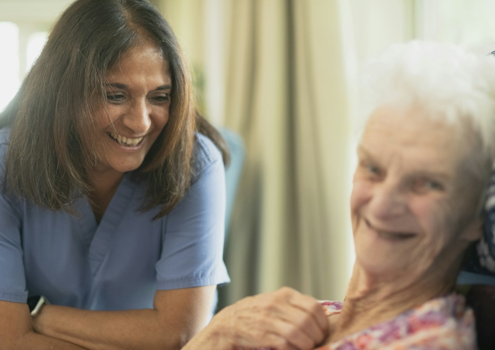 A smiling caregiver leans in close to an older woman, who is laughing with her.