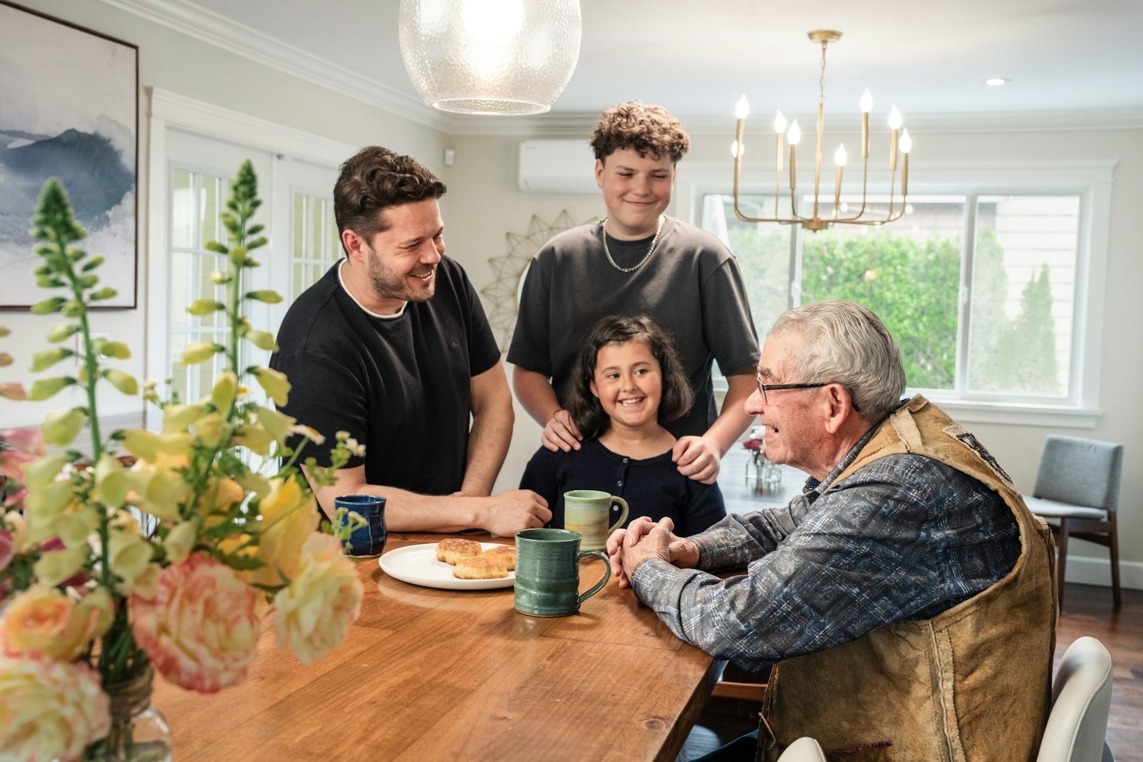 Three generations of a family gathered around a kitchen table with coffee, sharing a laugh.